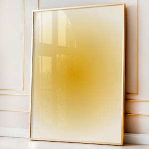 May include: A large, rectangular framed artwork with a gold-colored frame. The artwork features a gradient of yellow and cream colors. The frame is thin and has a reflective surface, and the artwork is leaning against a white wall.