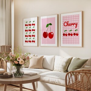May include: Three framed art prints featuring cherry designs. One print displays rows of red cherries with green stems on a white background. Another shows two cherries on a pink background. The third print has a pink gingham pattern with the word "Cherry" and cherries.