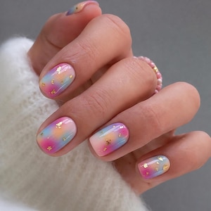 Puede incluir: Uñas cortas y redondeadas con un degradado suave de esmalte rosa pastel, naranja suave y azul claro. El diseño de uñas está acentuado con pequeños trozos dispersos de lámina de pan de oro metálico incrustados en la capa superior brillante.