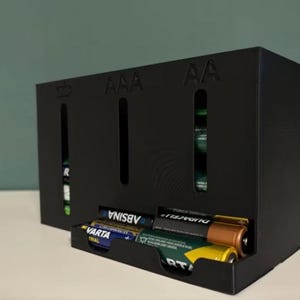 Könnte beinhalten: Ein schwarzer Batterieträger mit Schlitzen für AAA- und AA-Batterien. Der Träger hat eine Ablage mit verschiedenen AA-Batterien, darunter Marken von Varta und Duracell. Der Hintergrund ist hellgrün.