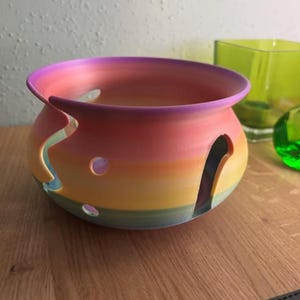 Puede incluir: Un cuenco de hilo de cerámica pintado a mano con un vibrante degradado de colores, incluyendo rosa, naranja, amarillo y azul. El cuenco tiene una abertura curva y varios diseños recortados. Un artículo decorativo para los entusiastas de la artesanía.