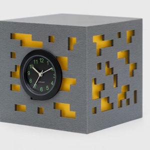 Può includere: Un orologio grigio a forma di cubo con quadrante nero e numeri verdi. Il cubo presenta ritagli pixelati che rivelano un interno giallo. L'orologio è un design moderno e geometrico.