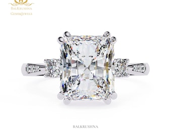 May include: A radiant-cut diamond engagement ring with a large, rectangular center stone and smaller round diamonds on the sides. The ring is set in a white metal band, and the design is elegant and classic. The brand name "BALKRUSHNA" is visible.