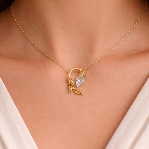 May include: A gold-colored necklace featuring a pendant with a clear, oval-shaped gemstone. The pendant is encircled by a gold-colored design with leaf accents. The necklace is worn on a person.