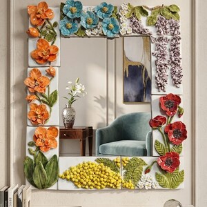 May include: A rectangular wall mirror framed by white ceramic tiles featuring three-dimensional floral reliefs. The frame displays orange orchids, blue blossoms, purple wisteria, yellow mimosa clusters, and red poppies with green leaves.