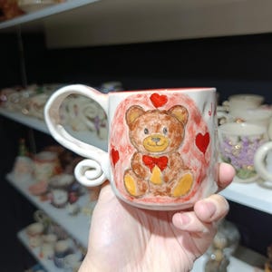 Puede incluir: Una taza de cerámica blanca con un diseño caprichoso de oso de peluche. El oso es marrón con una pajarita roja, rodeado de corazones rojos. La taza tiene un asa única con un remolino decorativo.