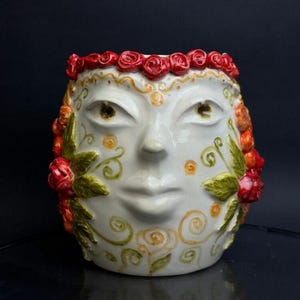 May include: A white ceramic face-shaped planter with red roses around the top and on the sides. The planter has a smiling face with detailed eyes and lips, and is decorated with gold and green floral designs.