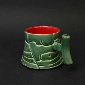 Peut inclure: Une tasse en céramique avec un extérieur vert et un intérieur rouge. La tasse a un design texturé unique avec une poignée sur le côté. La tasse est de couleur unie et semble être faite à la main.