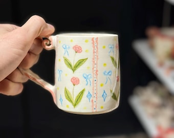 May include: A ceramic mug with a white base, adorned with hand-painted pink flowers, blue bows, and yellow dots. The mug features a curved handle and a decorative pink and white border. The design is whimsical and delicate.