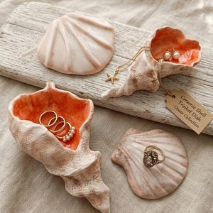 Może przedstawiać: Dwie ceramiczne miseczki w kształcie muszli o teksturowanej powierzchni zewnętrznej i gładkim, jaskrawopomarańczowym wnętrzu. Jedna miseczka jest otwarta i zawiera złote pierścionki, druga jest zamknięta. Etykieta głosi: Ceramic Shell Trinket Dish - Ocean Collection.