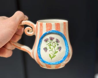 May include: A ceramic mug with a whimsical design. The mug features vertical orange and white stripes, a blue oval with a floral illustration, and a uniquely shaped handle. The mug is designed for hot beverages.