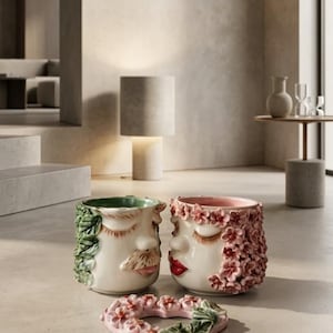 May include: Two ceramic face planters and a heart-shaped ornament. One planter features a moustache and green leaf hair, while the other has red lips and pink flower hair. The heart ornament is decorated with matching pink flowers and green leaves.