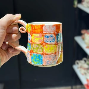 Pode incluir: Uma caneca de cerâmica colorida com uma alça única. A caneca apresenta um design de patchwork com quadrados laranja, rosa, amarelo, azul e verde. A alça é de cor rosa claro. A caneca é segurada por uma pessoa.