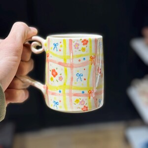 Pode incluir: Uma caneca de cerâmica pintada à mão com um design floral e laços. A caneca tem uma base creme com um padrão de xadrez rosa e amarelo. Pequenas flores e laços azuis decoram a superfície. A alça tem uma forma curva distinta.