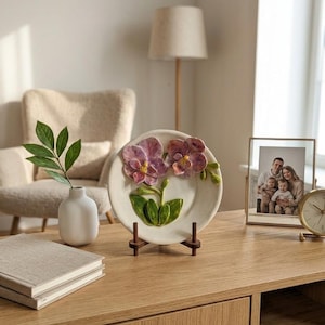 Può includere: Un piatto decorativo in ceramica con un design a orchidea 3D con accenti viola e verdi. Il piatto è esposto su un supporto in legno, un elemento decorativo floreale.