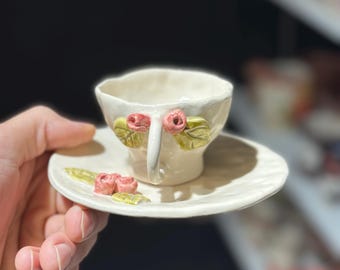 Puede incluir: Un juego de taza y platillo de cerámica hecho a mano. La taza blanquecina presenta un asa y está adornada con rosas rosas y hojas verdes. El platillo a juego también tiene detalles de rosas y hojas. Una adición encantadora.