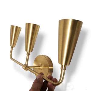 May include: A brass-colored wall sconce with three cone-shaped light fixtures. The sconce has a circular base and a curved arm design. The light fixtures are arranged in a horizontal line, with the largest fixture on the right.