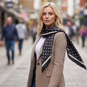May include: A woman wearing a brown and white houndstooth blazer, a white top, and a black scarf with white polka dots. The scarf has a white border. The background is a blurred street scene with pedestrians.