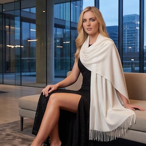 May include: A long, rectangular cream-colored shawl featuring a smooth, solid-colored fabric texture and delicate fringe detailing along the bottom edge. The shawl is draped over the shoulders and arms, showcasing its soft, lightweight appearance.