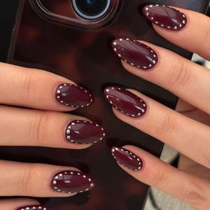 May include: Close-up of manicured nails painted a deep burgundy colour, adorned with small silver studs along the edges. The nails are oval-shaped and glossy, creating a stylish and elegant look. The background includes a tortoiseshell phone case.