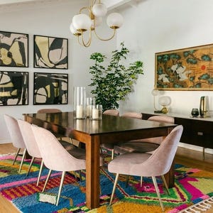 May include: A dining room with a dark wood table and pink chairs, set on a vibrant rug. The walls display abstract art and a decorative painting. A gold chandelier and a cabinet with decor complete the scene.