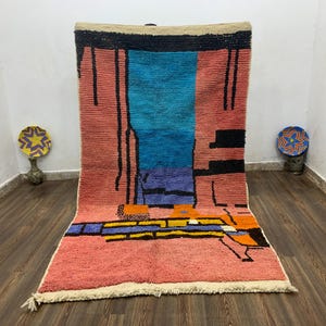 May include: A large, rectangular rug with a colorful abstract design. The rug features a salmon-colored field with a central panel of blue and purple. Black lines and geometric shapes add contrast. The rug has a cream-colored fringe.