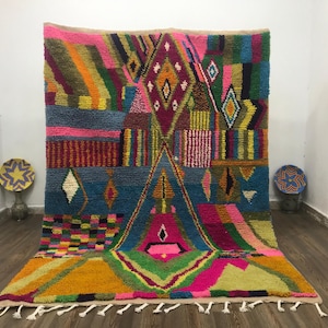 Pode incluir: Um vibrante tapete marroquino tecido à mão com um design geométrico. O tapete apresenta uma mistura de cores, incluindo rosa, azul, amarelo e verde. O design inclui formas de diamante e triângulo. O tapete tem uma franja ao longo da borda inferior.