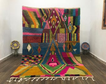 Puede incluir: Una vibrante alfombra marroquí tejida a mano con un diseño geométrico. La alfombra presenta una mezcla de colores que incluyen rosa, azul, amarillo y verde. El diseño incluye formas de diamante y triángulo. La alfombra tiene flecos en el borde inferior.