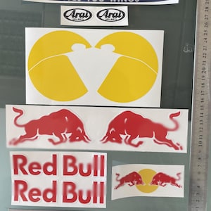 Peut inclure: Un ensemble d'autocollants comprenant une bande bleue avec le texte GIVES YOU WINGS, deux logos Arai Helmet, deux grands cercles jaunes, deux silhouettes de taureaux rouges, deux étiquettes Red Bull et un petit autocollant taureau et cercle.