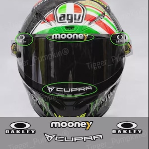 Op de afbeelding: Zwarte motorhelm met sponsorstickers. De helm toont de tekst "VISOR SPONSOR DECALS / STICKERS FOR MARCO BEZZECCHI HELMET". Logo's zijn onder meer "AGV", "mooney", "CUPRA" en "OAKLEY".