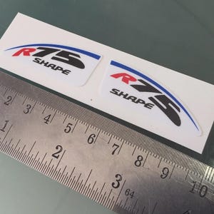 Op de afbeelding: Twee witte stickers met de tekst "R75 SHAPE" in rood, zwart en blauw. Een metalen liniaal is naast de stickers geplaatst. De stickers zijn waarschijnlijk voor een voertuig of product.