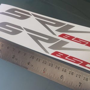 May include: Two gray and red SRV 850 decals on a white sheet, with a ruler for scale. The decals have a stylized design. The ruler shows measurements in inches.