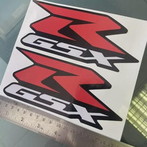 Puede incluir: Dos calcomanías rojas y negras de motocicleta GSX con letras blancas. Las calcomanías están sobre un fondo blanco, y una regla de metal se coloca en la parte inferior de la imagen, midiendo en centímetros.