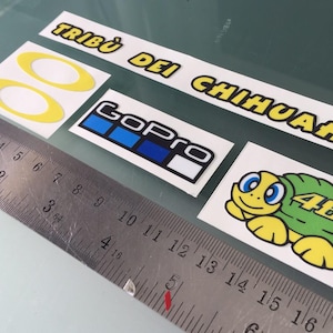 Peut inclure: Un ensemble de quatre autocollants comprenant un huit jaune, un logo GoPro bleu et blanc, une tortue verte avec le numéro 46, et une longue bande portant l'inscription TRIBÙ DEI CHIHUAHUA, à côté d'une règle métallique graduée jusqu'à 16 centimètres.