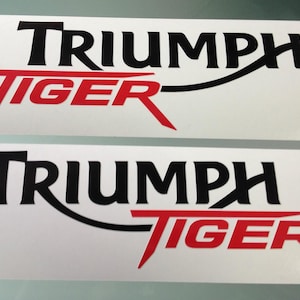 Puede incluir: Dos pegatinas rectangulares que muestran la palabra TRIUMPH en letras mayúsculas negras y la palabra TIGER en letras mayúsculas rojas. El texto está dispuesto en un diseño gráfico estilizado con una línea negra curva que conecta ambas palabras.
