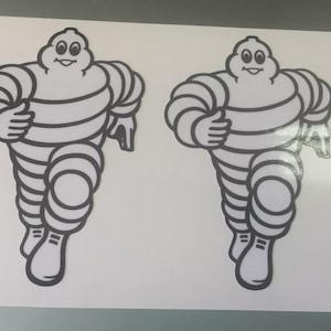 Könnte beinhalten: Zwei weiße Aufkleber mit dem Michelin-Männchen, einer Zeichentrickfigur aus weißen Reifen. Das Michelin-Männchen wird in einer Laufpose mit schwarzen Umrissen dargestellt. Ein Lineal befindet sich neben den Aufklebern zur Maßstabsbestimmung.