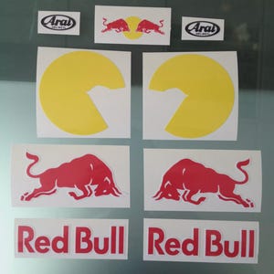 Könnte beinhalten: Eine Sammlung von Aufklebern mit dem Red Bull-Logo, einschließlich der Worte "Red Bull" in roten Blockbuchstaben, einer roten Bullensilhouette und gelben Kreismotiven. Ebenfalls enthalten sind Arai Helmet-Aufkleber.