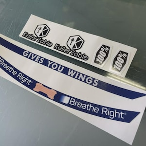 Può includere: Una collezione di adesivi bianchi con testo nero e blu. Gli adesivi includono le frasi "Gives You Wings" e "Breathe Right", insieme ai loghi "KellerZable" e "100%". L'adesivo "Breathe Right" ha una grafica a benda beige.