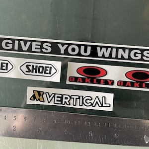 Könnte beinhalten: Eine Sammlung von Aufklebern, darunter ein langer schwarzer Streifen mit dem weißen Text GIVES YOU WINGS, zwei SHOEI-Logos, zwei OAKLEY-Logos mit roten Ovalen und ein VERTICAL-Logo, platziert über einem Metalllineal mit einer Länge von 21 cm.