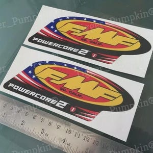 May include: Two FMF Powercore 2 stickers. Each sticker has a yellow and red design with the text "FMF" and "POWERCORE 2 SO. CALIF." The stickers have a patriotic red, white, and blue color scheme.