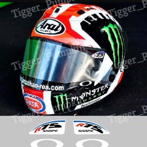 Peut inclure: Un casque de moto avec un design graphique rouge, blanc et noir, incluant les logos Monster Energy et Arai. La visière porte le texte jonathan-rea.com. En dessous, deux autocollants R75 SHAPE et deux ovales blancs sont présentés.