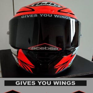 May include: A motorcycle helmet featuring a bright red and black colour scheme with a dark tinted visor. The text GIVES YOU WINGS is printed on the visor. Additional decals include the text acebsa in red diamonds and HJC logos on the helmet.