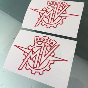 Puede incluir: Dos pegatinas rectangulares blancas con un logotipo rojo de MV Agusta. El diseño consiste en un emblema en forma de engranaje con las letras MV en el centro y la palabra AGUSTA impresa en una pancarta en la parte superior del gráfico.