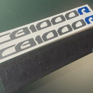 以下が含まれることがあります： グレーとブルーのC1000Rバイクデカール2枚。デカールは白い背景にあり、隣に黒い定規があります。デカールの長さは約15cmです。