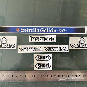 Op de afbeelding: Een verzameling diverse stickers, waaronder een lange blauwe Estrella Galicia 0.0-strip, twee Verisure-logo's, een Insta360-label, twee Vertical-tekststickers en twee Shoei-emblemen, gerangschikt boven een metalen liniaal van 26 cm lang.