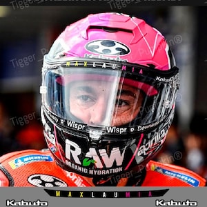 Peut inclure: Un casque de moto rose et noir avec des autocollants de sponsors sur la visière pour Aleix Espargaro. La visière affiche des textes comme MAXLAUMIA, Wispr et RAW ORGANIC HYDRATION. Le casque porte la marque Kabuto et divers logos de sponsors.