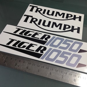 Könnte beinhalten: Zwei Sätze Aufkleber auf weißem Trägerpapier. Ein Satz zeigt den schwarzen Schriftzug TRIUMPH. Der andere Satz zeigt den schwarzen Schriftzug TIGER gefolgt von dem grauen Text 1050. Ein Metalllineal zeigt eine Länge von 20 cm.