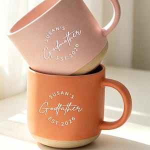 Puede incluir: Dos tazas de cerámica apiladas con acabado mate. La taza superior es de color rosa claro con el texto blanco "Susan's Godmother EST. 2026". La taza inferior es de color naranja terracota con el texto blanco "Susan's Godfather EST. 2026".