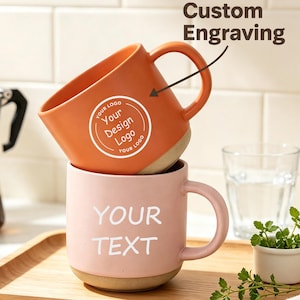 Op de afbeelding: Twee keramische mokken verticaal op elkaar gestapeld. De bovenste mok is oranje met een cirkelvormig logo en de tekst "Your Design Logo" en "Custom Engraving" met een pijl. De onderste mok is lichtroze met de witte tekst "YOUR TEXT" erop.