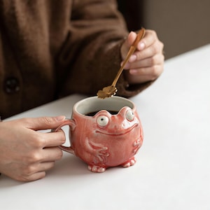 May include: A whimsical, coral-colored ceramic mug shaped like a frog. The mug features a detailed frog face with bulging eyes and small arms and legs. A gold-colored spoon with a flower-shaped bowl rests above the mug.